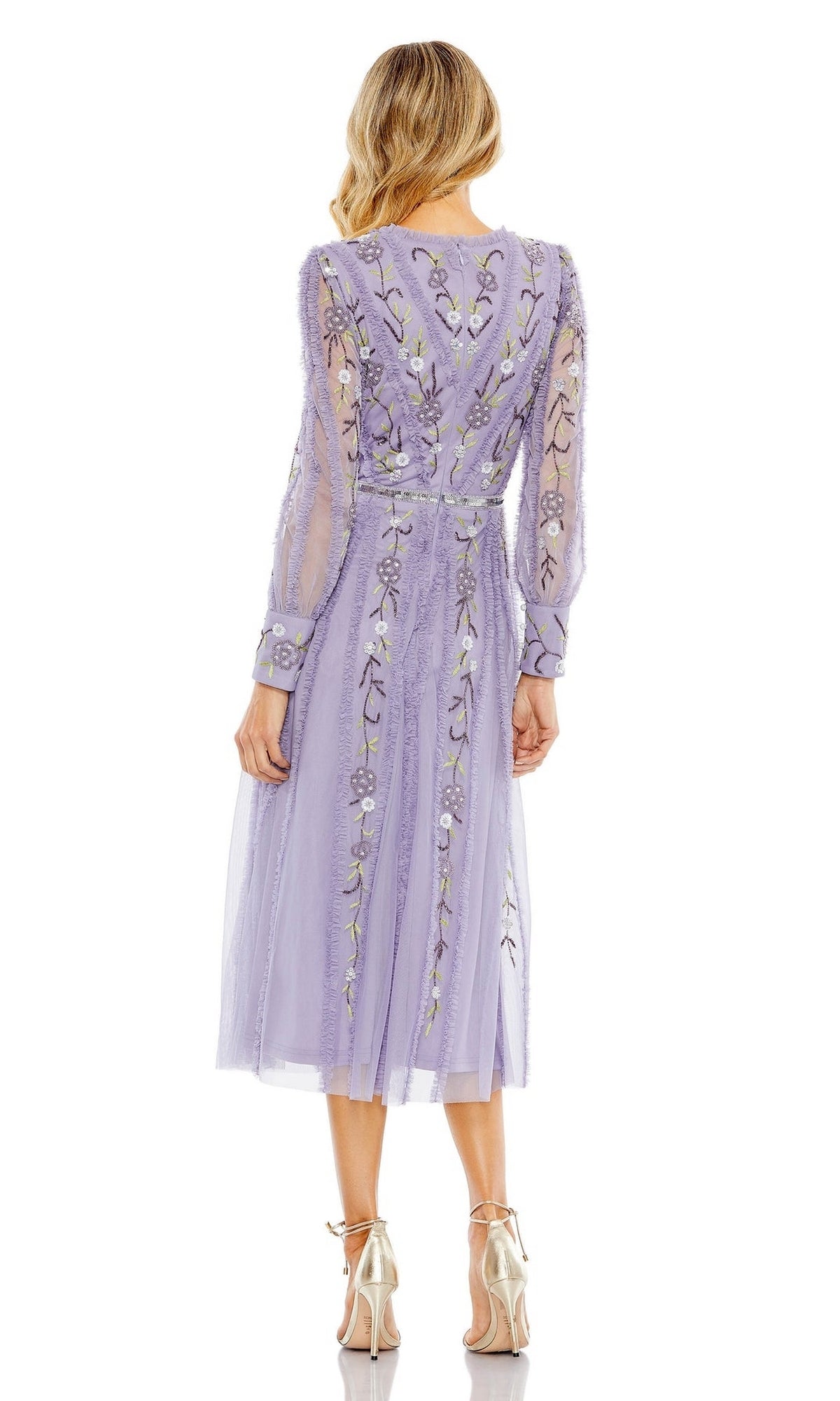 A woman stands facing away, wearing the Knee Length Wedding Guest Dress 9133 by Mac Duggal in lavender with floral embroidery and sheer fabric, paired with metallic high-heeled shoes.